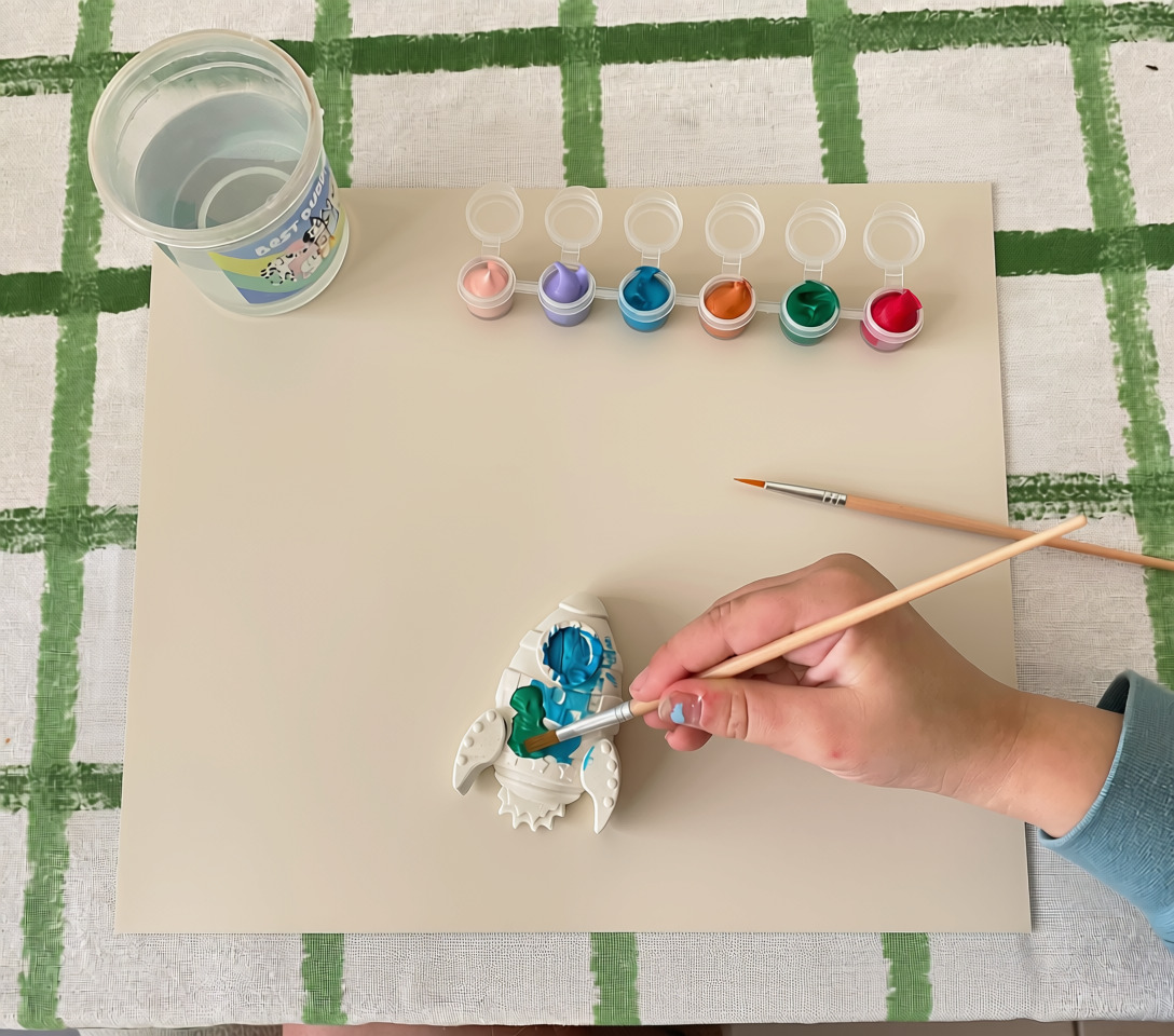 Child painting a plaster rocket in blue and green at a craft table